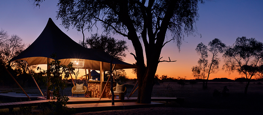 Luxury tent at Tuludi Camp, Botswana, featuring a private seating area under a large canopy, surrounded by trees, with a vibrant sunset sky in the background, highlighting the camp's refined safari experience.