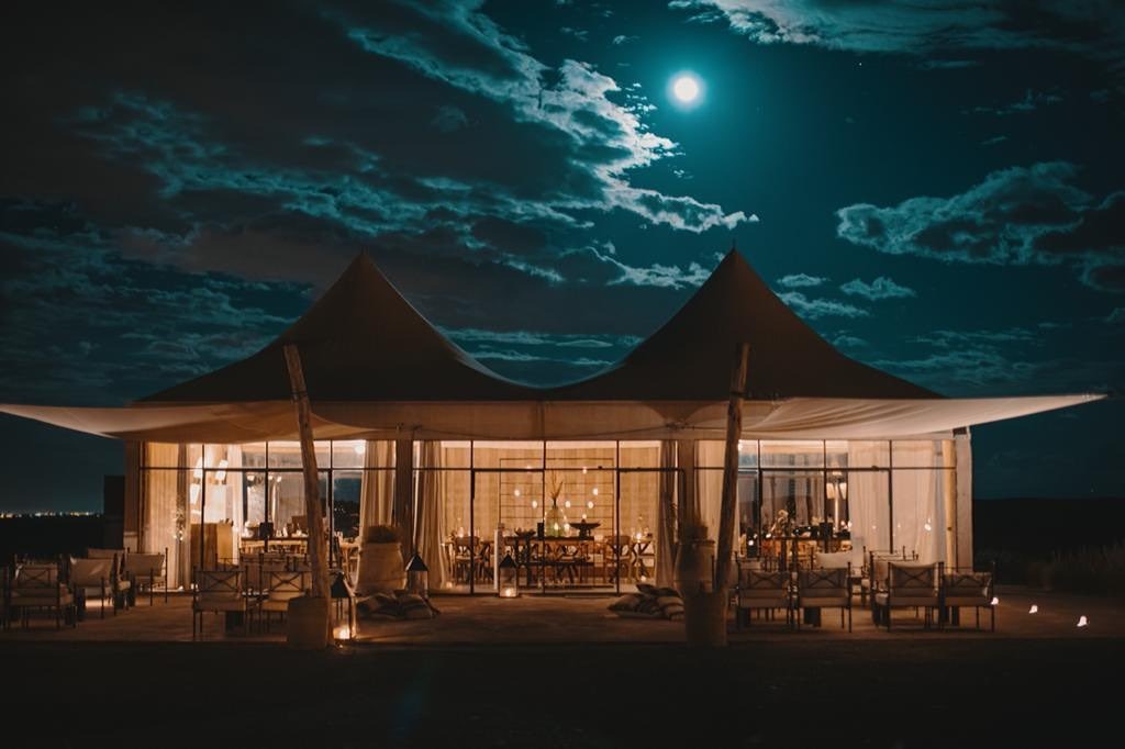 A twin-peaked glass pavilion glows against a moonlit desert sky, clouds drifting past a near-full moon.