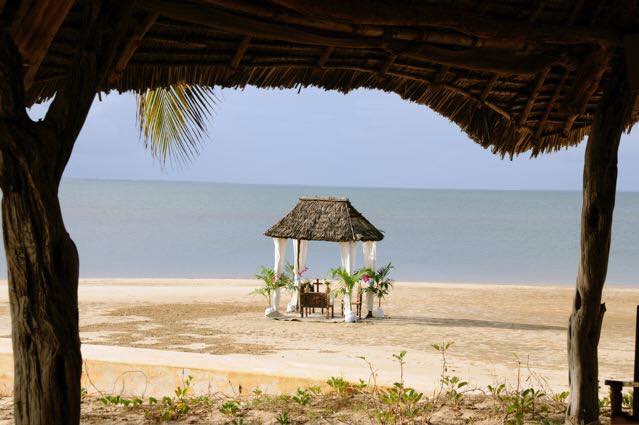 Thatched beach gazebo hung with white curtains and set with chairs on the sand, framed by a thatched shelter at Manda Bay Lodge, Kenya.