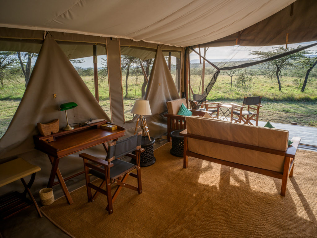 Superior tent interior with a writing desk and green banker's lamp, director's chair and sofa facing an open front onto acacia grassland.