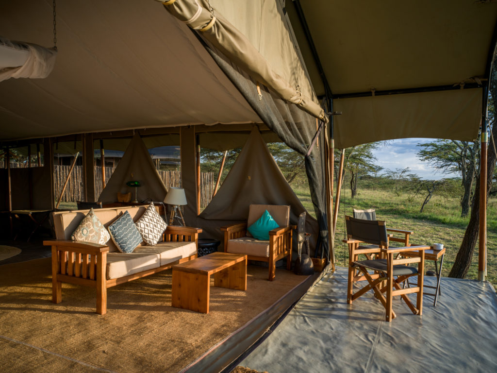 Superior tent lounge in late afternoon sun, with a wooden sofa and armchair inside and director's chairs on the veranda facing the bush, Kenya.