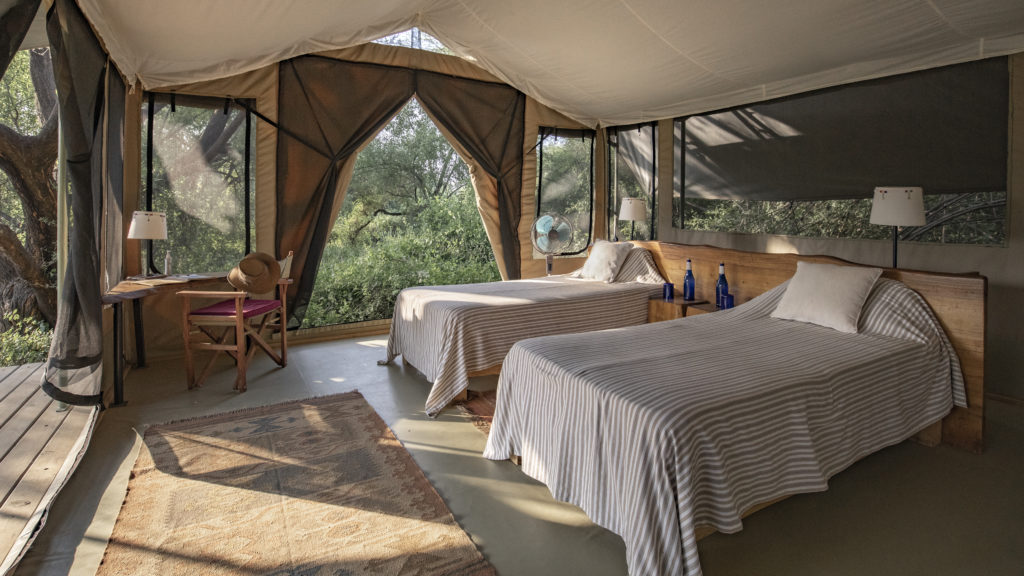 Twin tent at Shompole Wilderness with striped bedspreads, a timber headboard, blue glass bottles and a writing desk facing green bush.