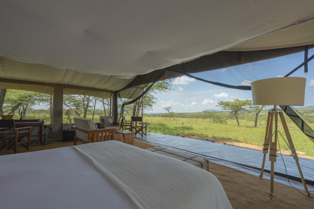 View from the bed across a superior tent at Kandili Camp, with a tripod floor lamp, sitting area and open sides onto green Maasai Mara plains.