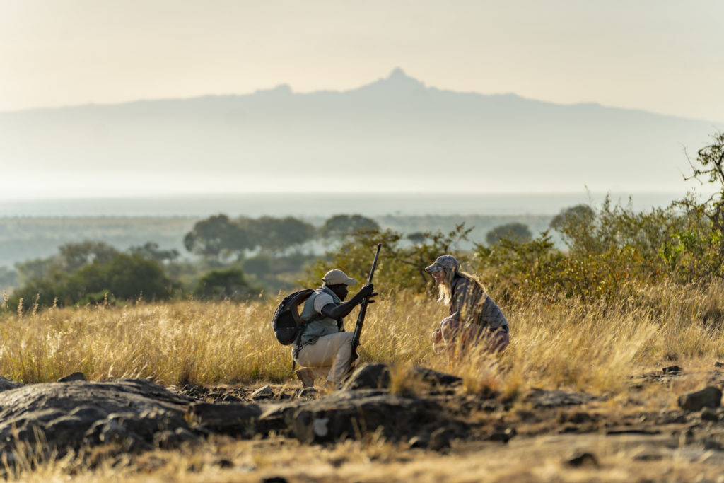Armed walking guide and guest crouched in long grass on a bush walk, with Mount Kenya rising through the haze behind.