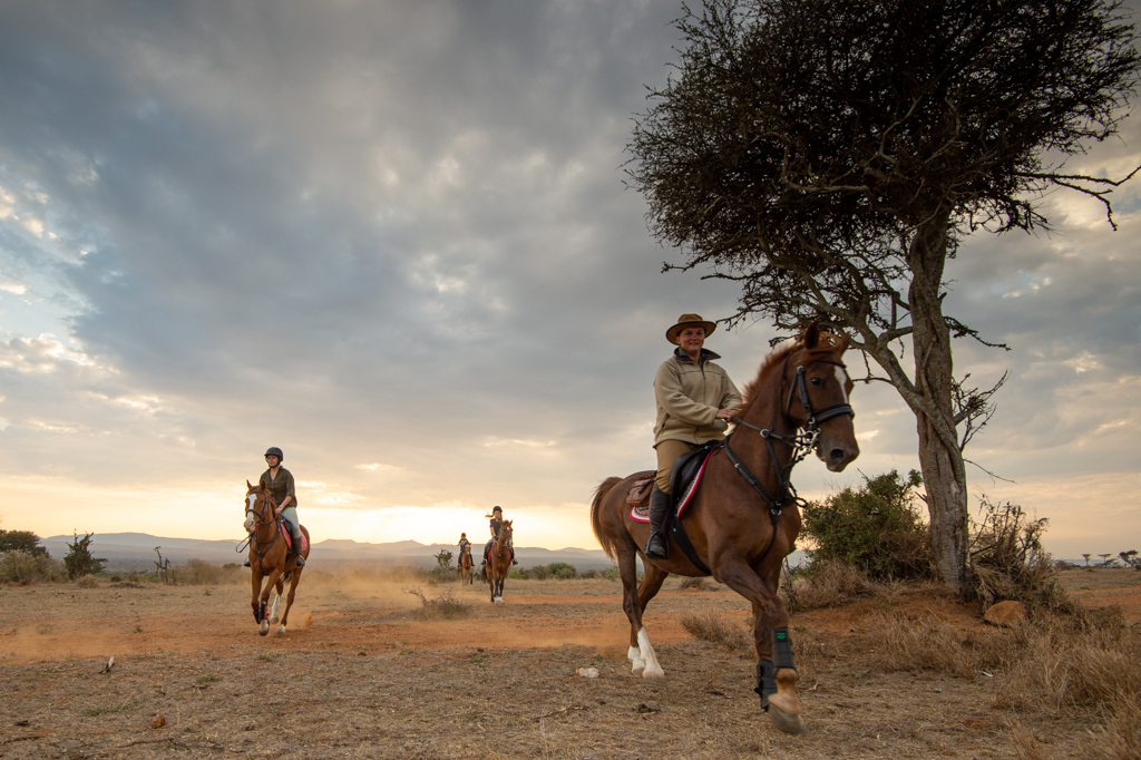 Riders on horseback trotting across the dusty Laikipia plains beneath a stormy sky on an El Karama riding safari.