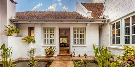 The whitewashed inner courtyard entrance of Hamilton House, a stepping-stone path crossing a small koi pond bordered by tropical plantings, carved wooden sculptures and sage-green French doors