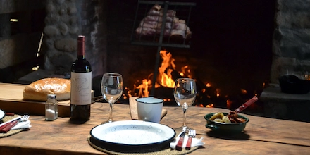 A candlelit Patagonian asado set beside an open fire, with local wine, simple tableware and the warm glow of flames reflecting the rhythm of camp life.