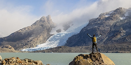 A traveller stands triumphantly on a rocky outcrop facing a sweeping glacier that spills into a turquoise Patagonian lake.