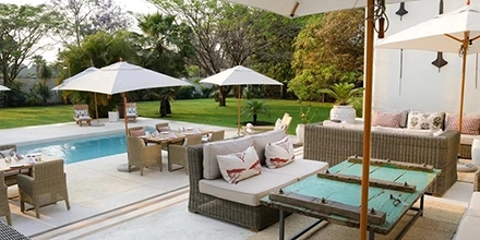 The poolside lounge terrace at Latitude 15° in Lusaka, with wicker sofas, printed cushions, a weathered turquoise coffee table and white parasols set against a lawn and mature tree canopy.