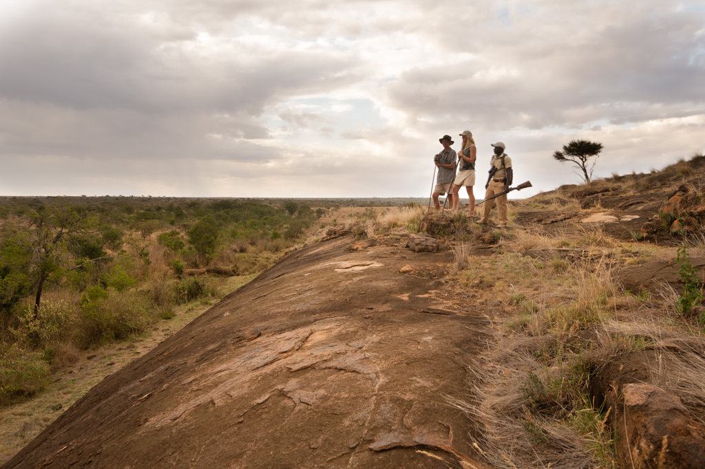 Two walkers and an armed guide standing on a rocky granite outcrop overlooking the Laikipia bush under a cloudy sky.