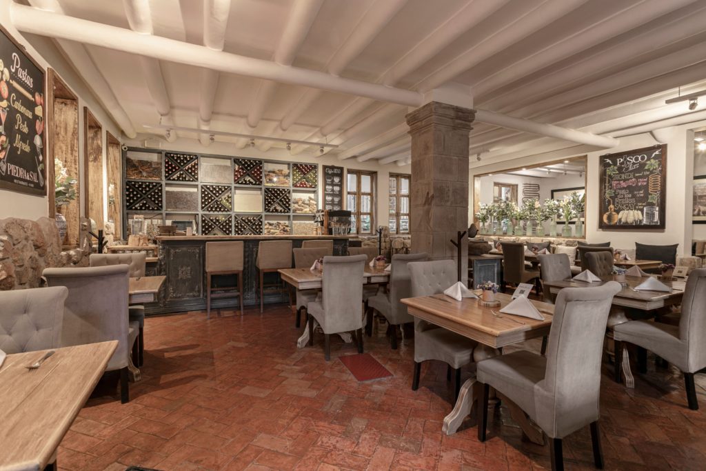 The dining room at Piedra & Sal, with upholstered chairs, wood tables set for service, and a wine wall behind the bar.