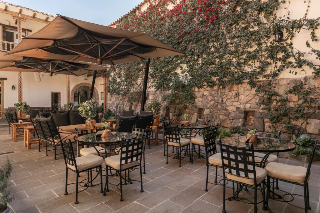 Wrought-iron café tables cluster beneath awning umbrellas as bougainvillea climbs the stone courtyard wall in late afternoon light.