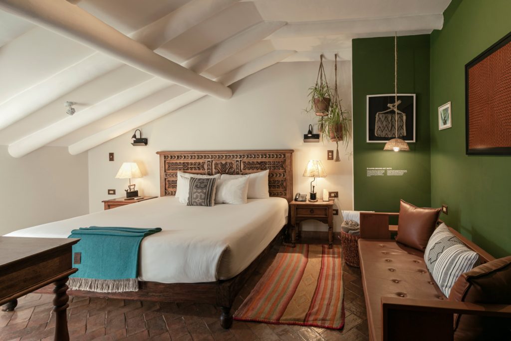 A Xapiri Ground Art Suite with a carved wooden headboard, teal throw, and a green feature wall displaying curated Amazonian textiles and artefacts.