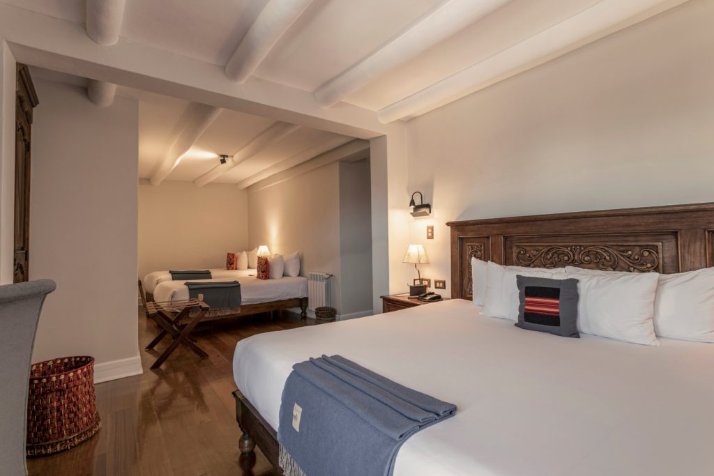 A suite with a carved wooden headboard, layered white bedding, and a second sleeping area visible through an archway, all under exposed white beams.