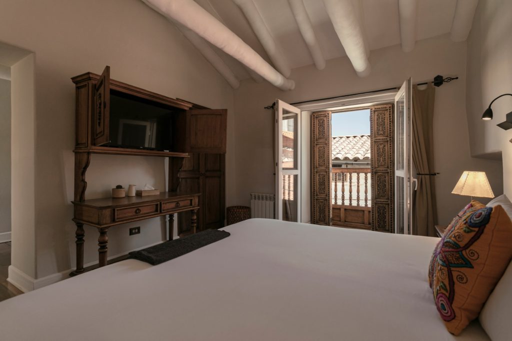 A suite bedroom with hand-carved armoire and desk, sloped beamed ceiling, and shuttered French doors opening onto a carved wooden balcony over Cusco's tiled rooftops.