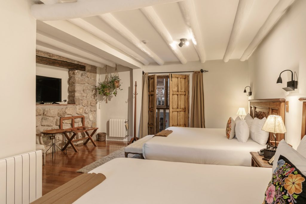 A twin-bedded suite with hand-carved wooden headboards, stone accent wall, and open shuttered doors leading onto a colonial balcony.