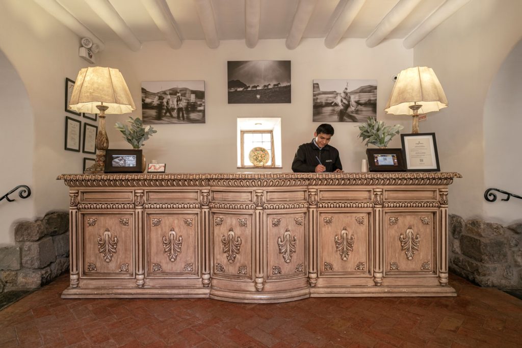 A carved wooden reception desk beneath framed black-and-white photography, with a staff member attending to guest arrivals.