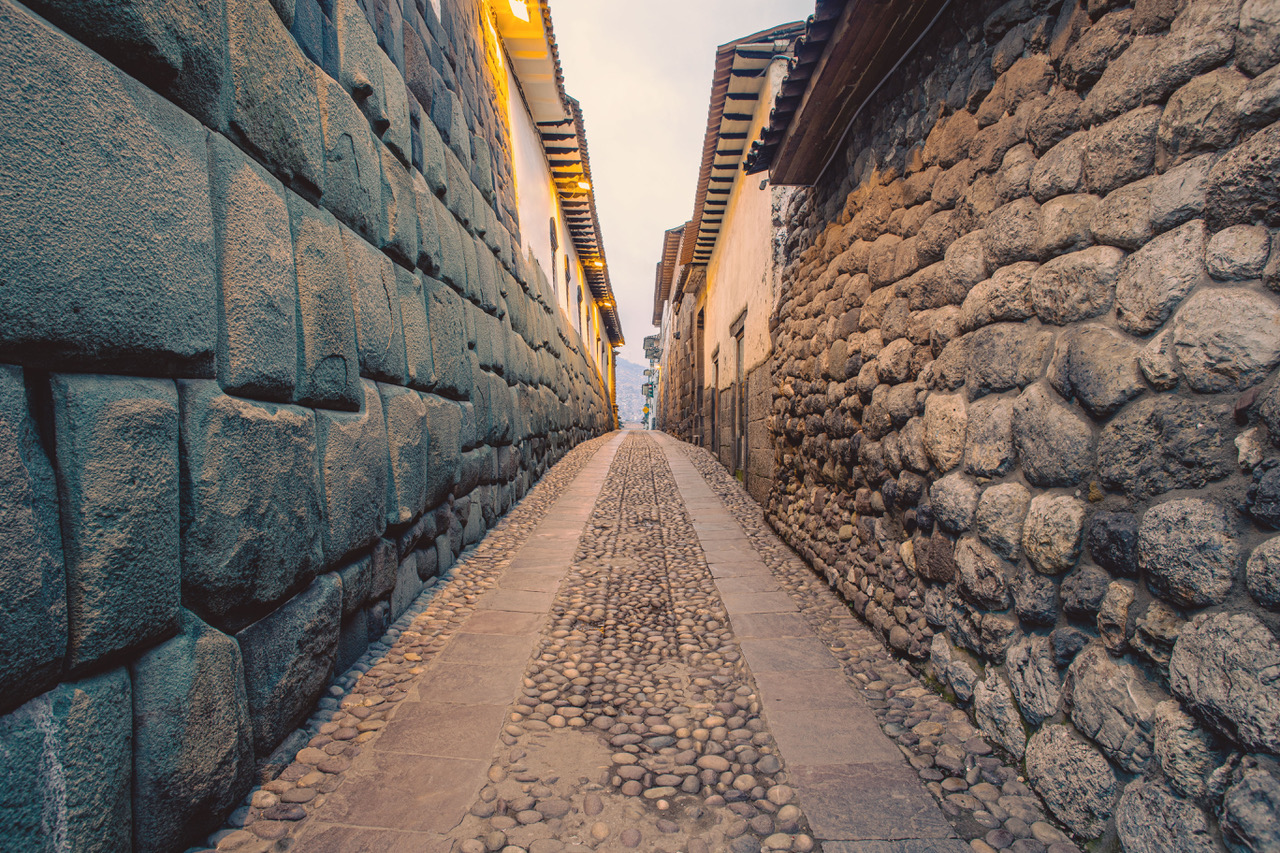 A cobbled Cusco laneway flanked by massive Incan polygonal stonework on one side and colonial rubble-stone walls on the other, leading toward the city beyond.