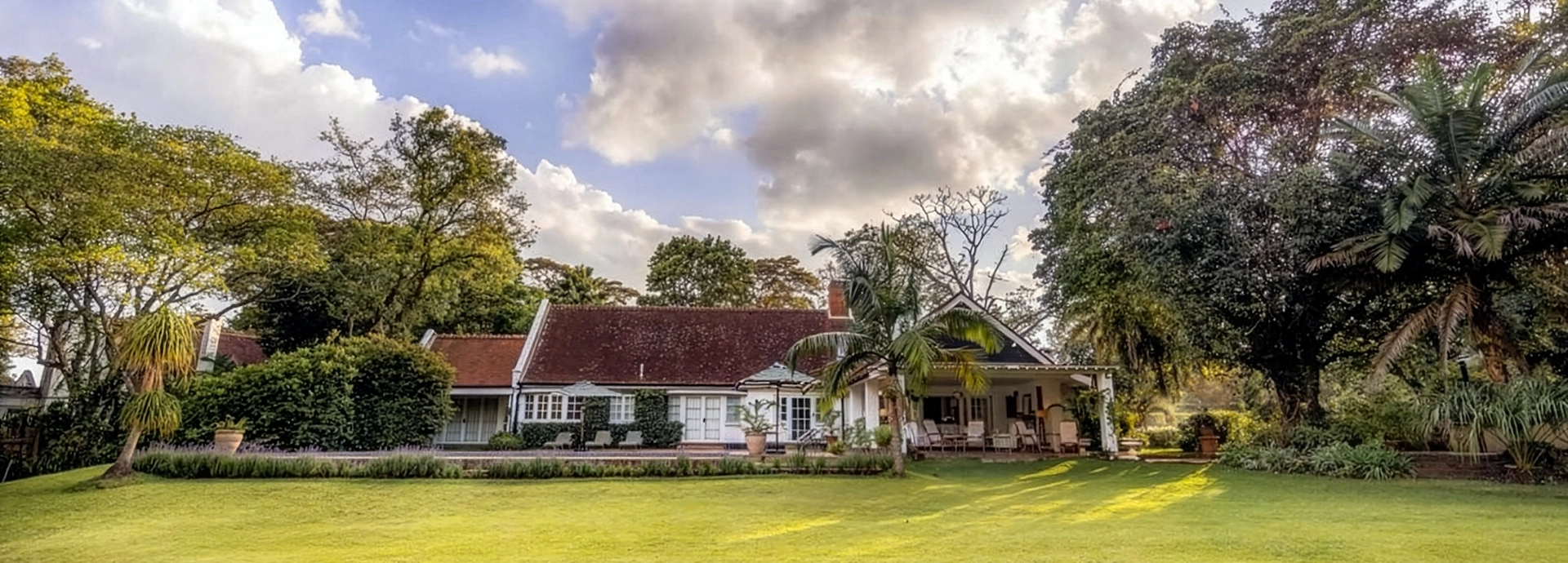 Hamilton House seen across its expansive manicured lawn at dusk, the long whitewashed 1930s facade with terracotta-tiled roof nestled among towering indigenous trees under a dramatic evening sky