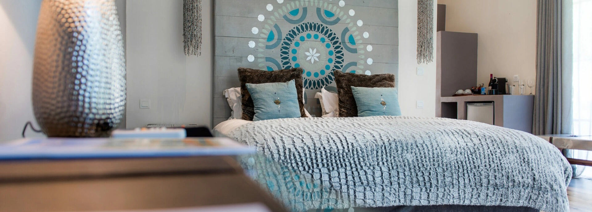 A guest room at Latitude 15° in Lusaka featuring a king bed dressed in a textured grey bedspread and teal and brown cushions, with a bespoke mandala artwork painted across the timber headboard panel.