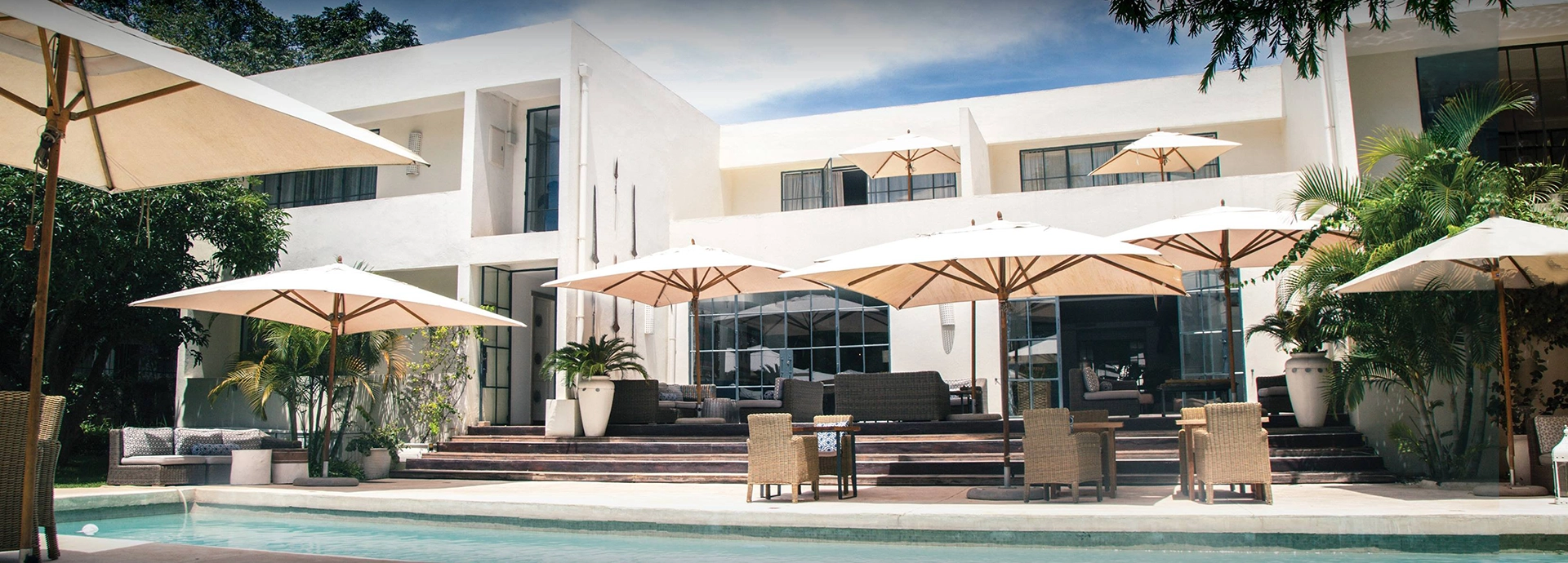 The pool terrace at Latitude 15° in Lusaka, with wicker furniture and large parasols arranged before a white modernist building framed by tropical palms and steel-framed glass windows.