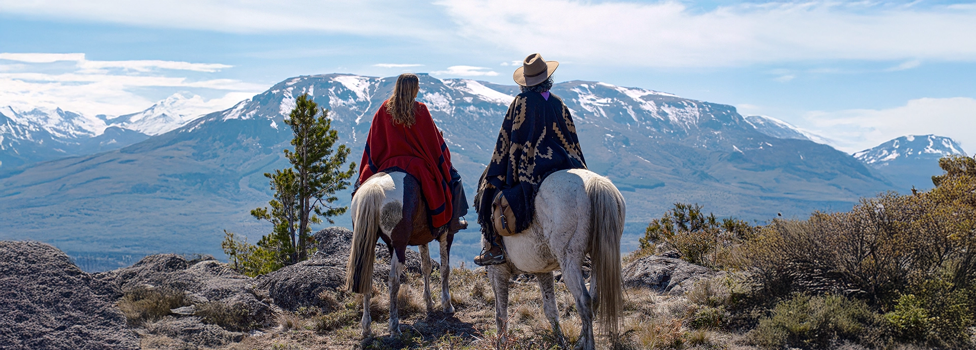 Two riders pause on horseback at a rocky viewpoint, gazing across rolling Patagonian valleys towards distant volcanic peaks.