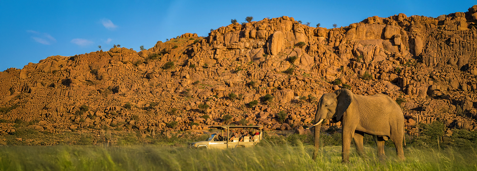 A game vehicle with tourists watches an elephant with a rocky hill in the background