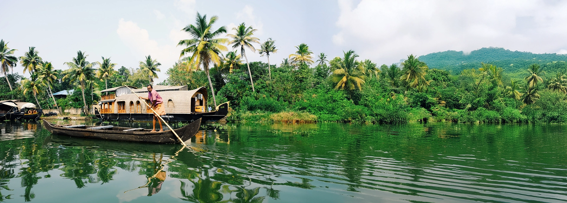 A traditional wooden canoe glides through Kerala&rsquo;s backwaters, with palm-lined banks and a Kettuvallam reflecting on the still water.