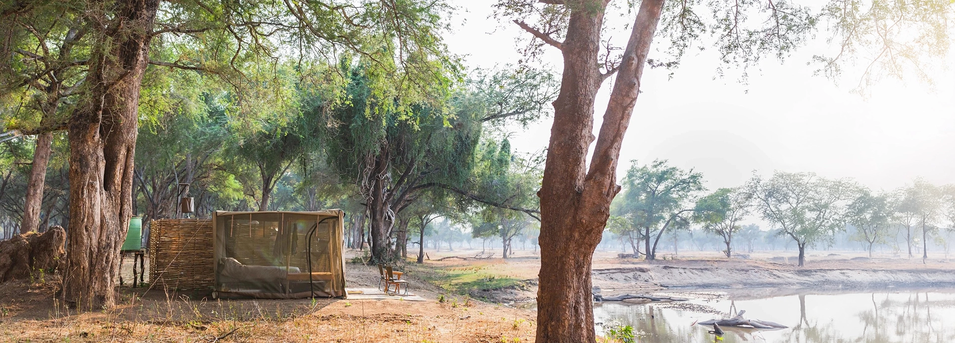 Simple safari tent set under trees beside a lagoon at Kutali Camp in the Lower Zambezi.