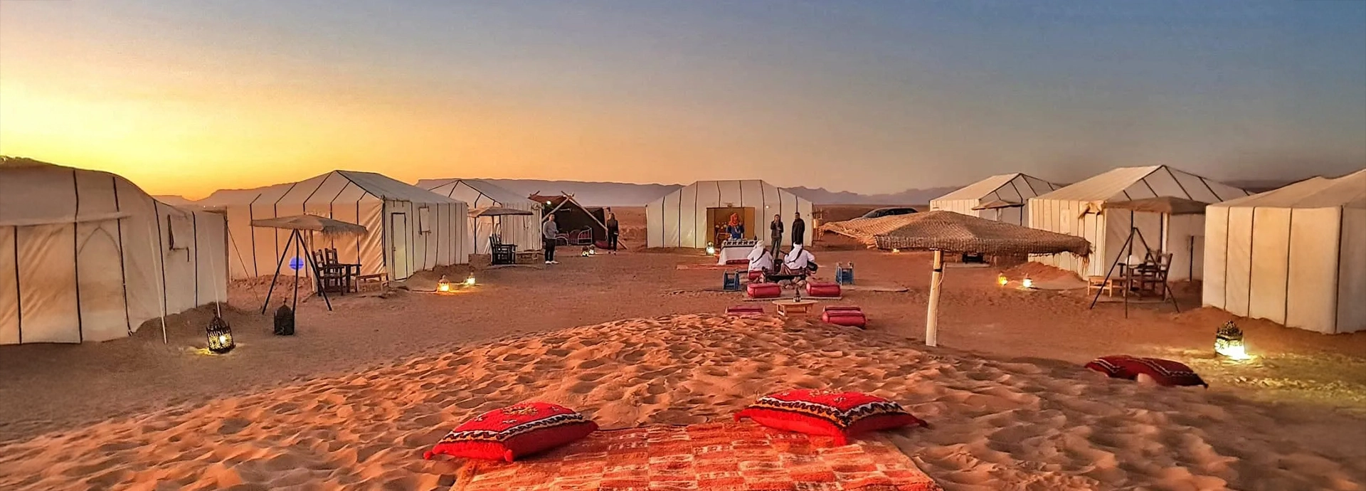 Luxury desert camp with glowing lanterns at sunset surrounded by sandy dunes in Merzouga.