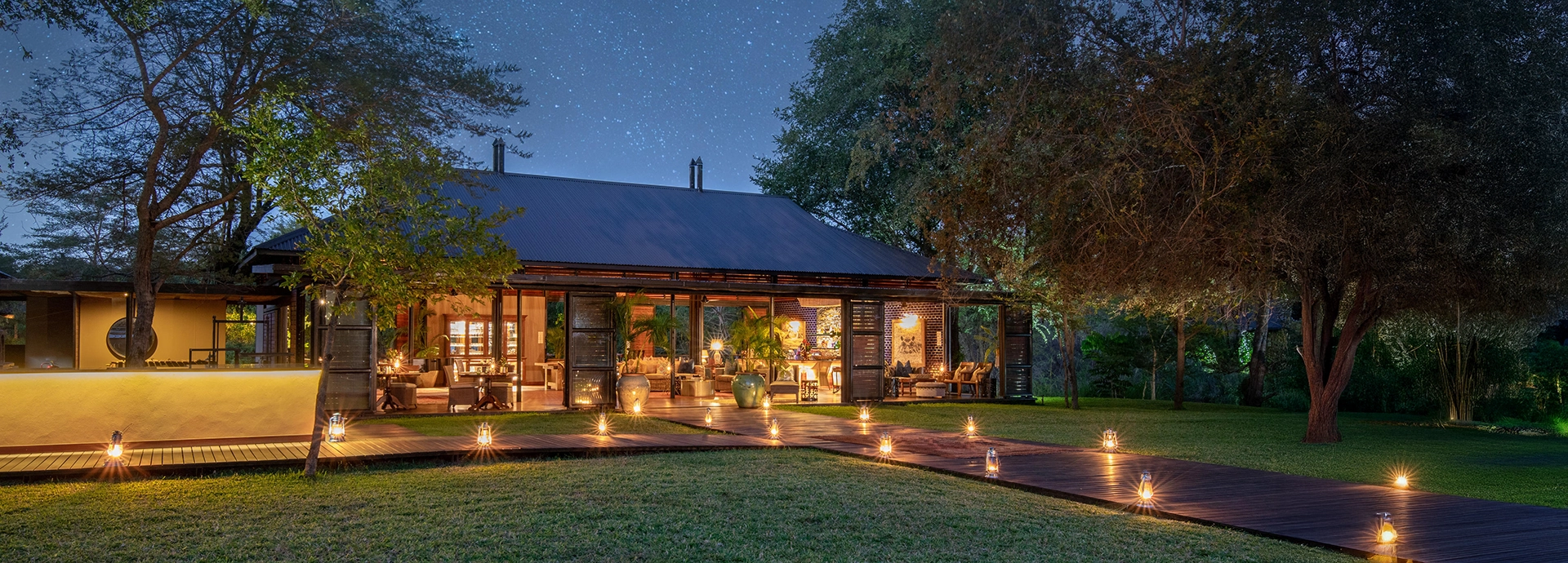 An evening view of Mukwa River Lodge shows the softly illuminated central areas set among mature trees beside the Zambezi River.