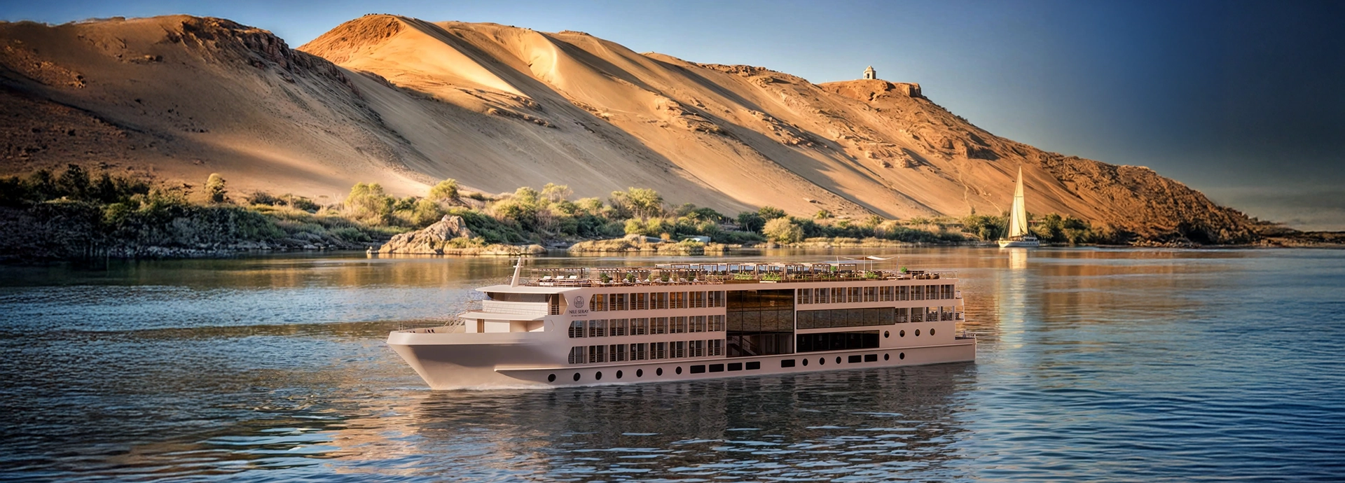 A&K Sanctuary Nile Seray gliding along the Nile at sunset, framed by sweeping sand dunes and the soft glow of late-afternoon light.