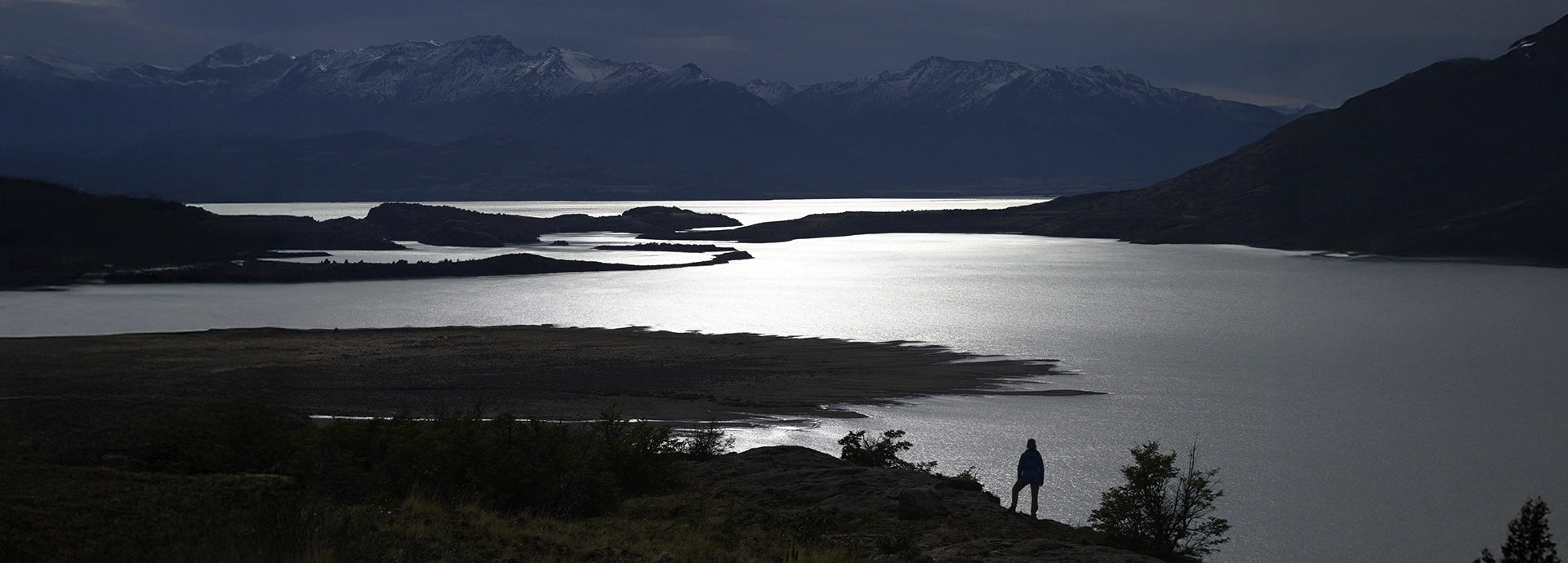 Person standing on a rocky outcrop gazing over the silver-lit waters of Lago San Martín at dusk.