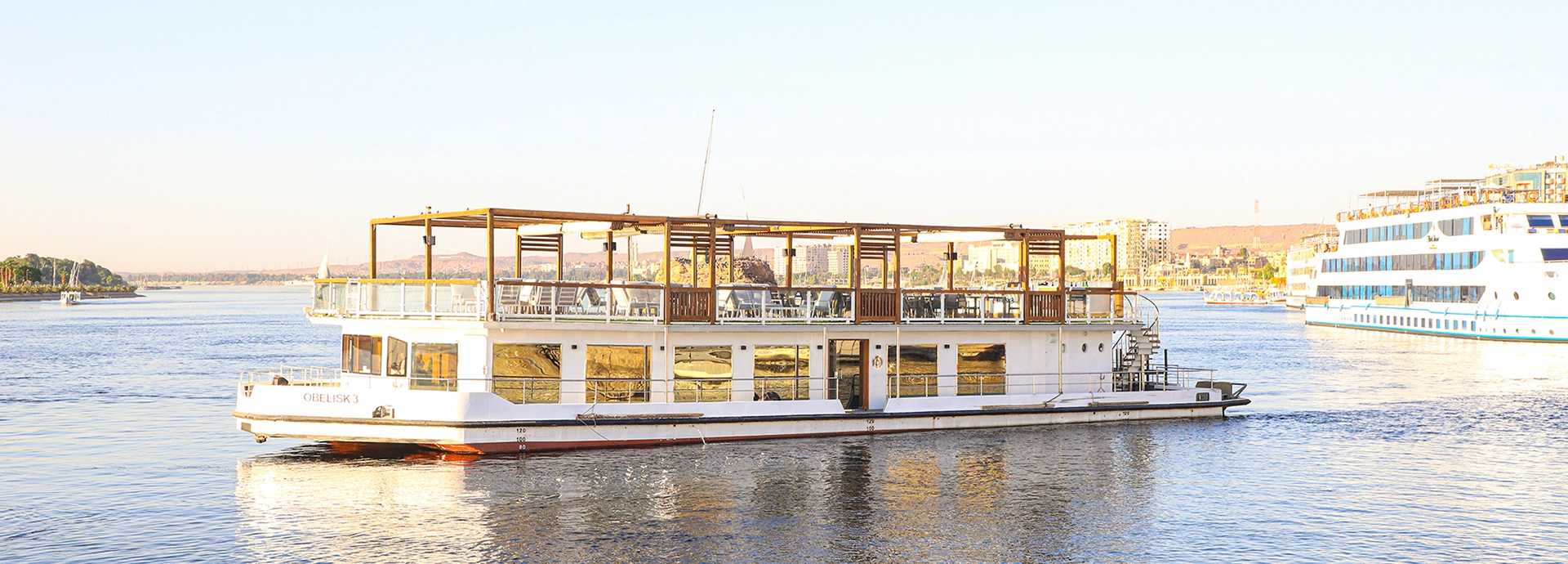 A sunlit Nile cruiser gliding along the river with an open upper deck and reflections shimmering across its windows.