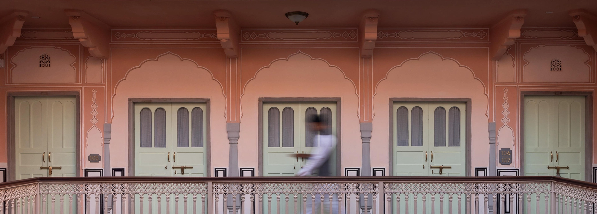 A corridor of pastel pink walls and mint green doors, with a blurred figure walking past ornate arches.