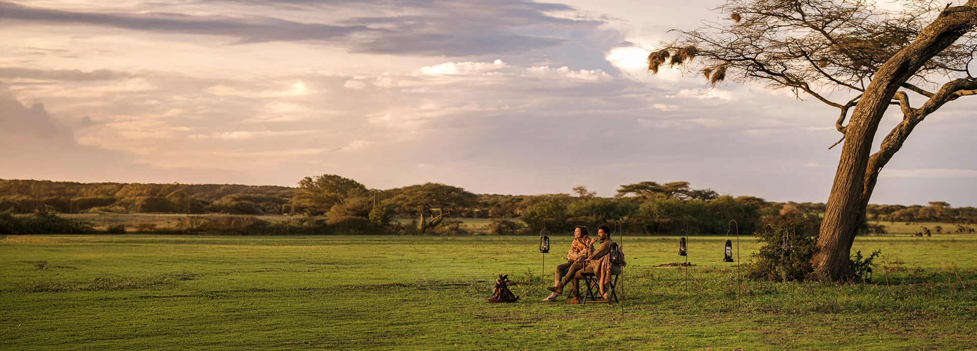 Guests enjoy a classic Serengeti sundowner beside the campfire, overlooking sweeping green plains and acacia trees as golden light settles over the migrating wilderness.