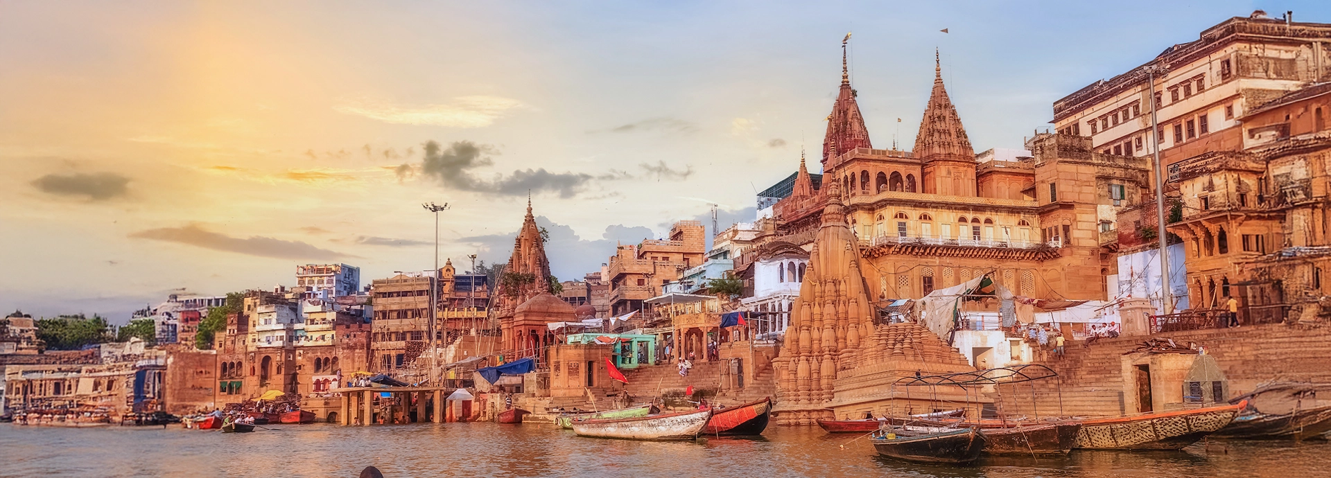 Golden-hued ghats of Varanasi at sunset with ornate temples and colourful buildings reflected in the Ganges.