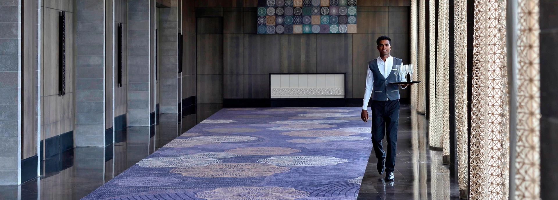A hotel staff member walks through a grand, softly lit corridor at Weligama Bay Marriott Resort & Spa carrying a tray of glasses.