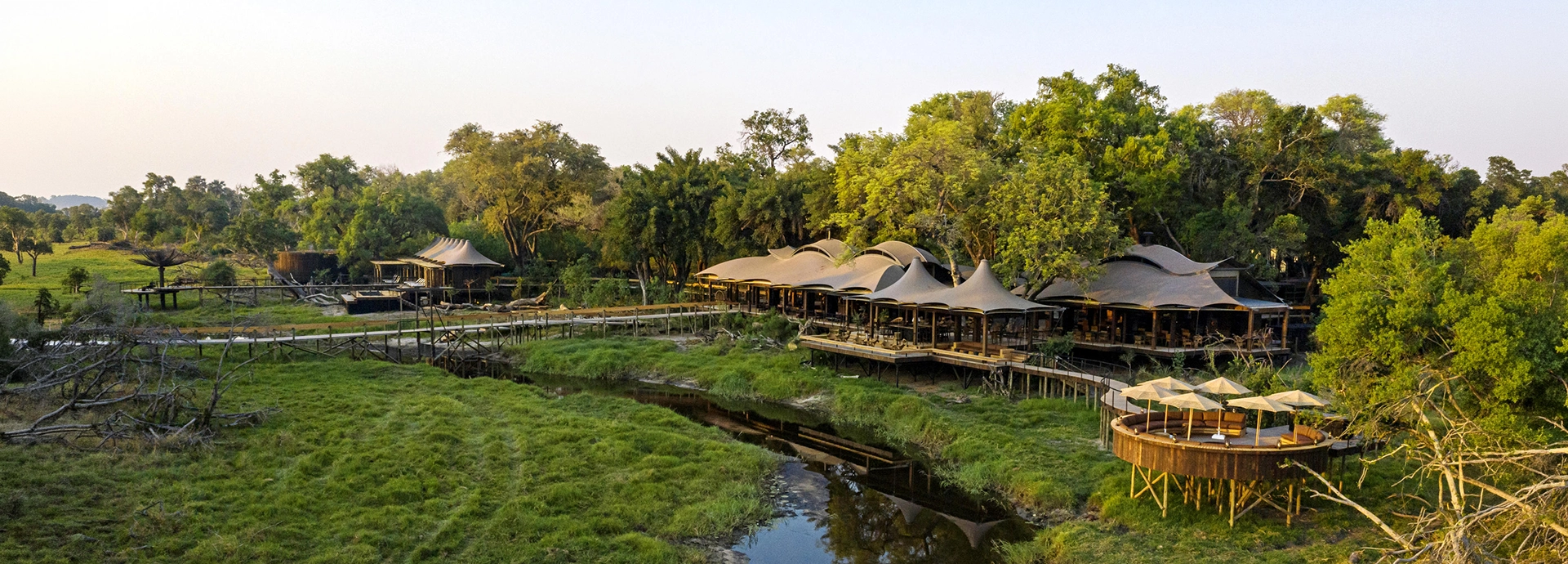 An aerial view of Xigera Safari Lodge's wave-shaped canvas roofline rising above the tree canopy, with elevated timber walkways extending over the lush green channels of the Okavango Delta at dusk.
