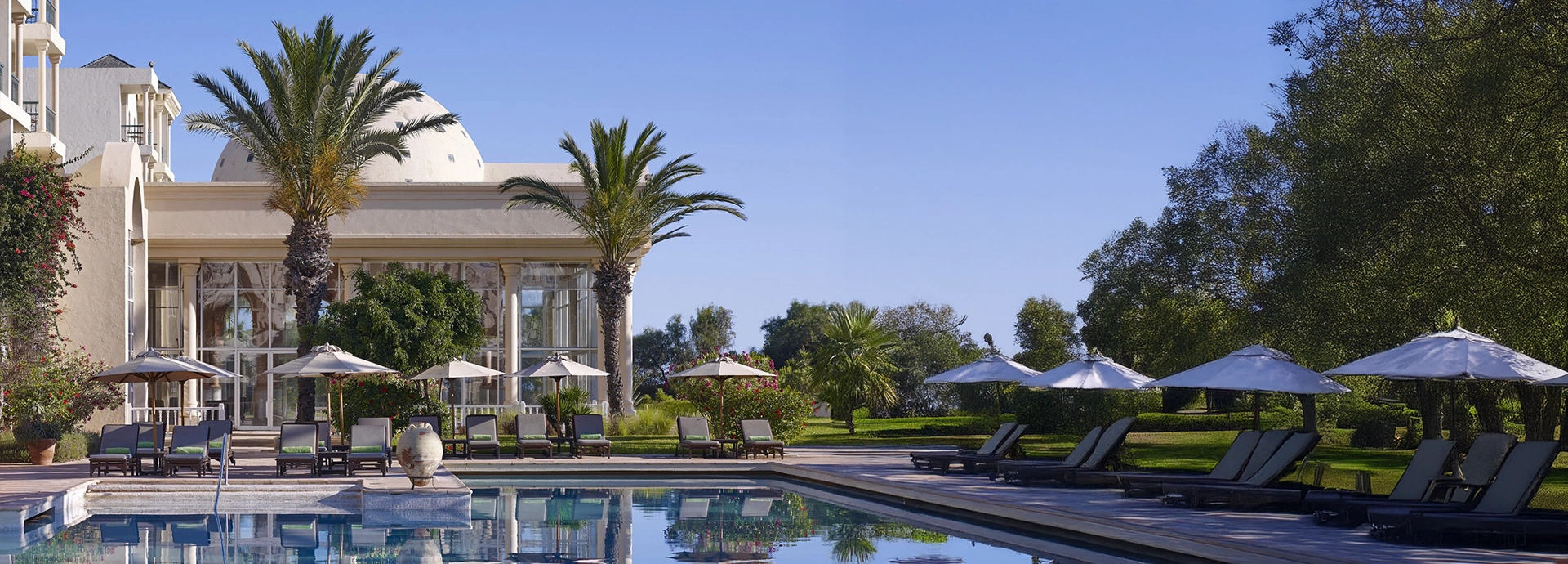 Poolside view at a Tunisian resort with sun loungers, parasols, and towering palms beside a domed white building.