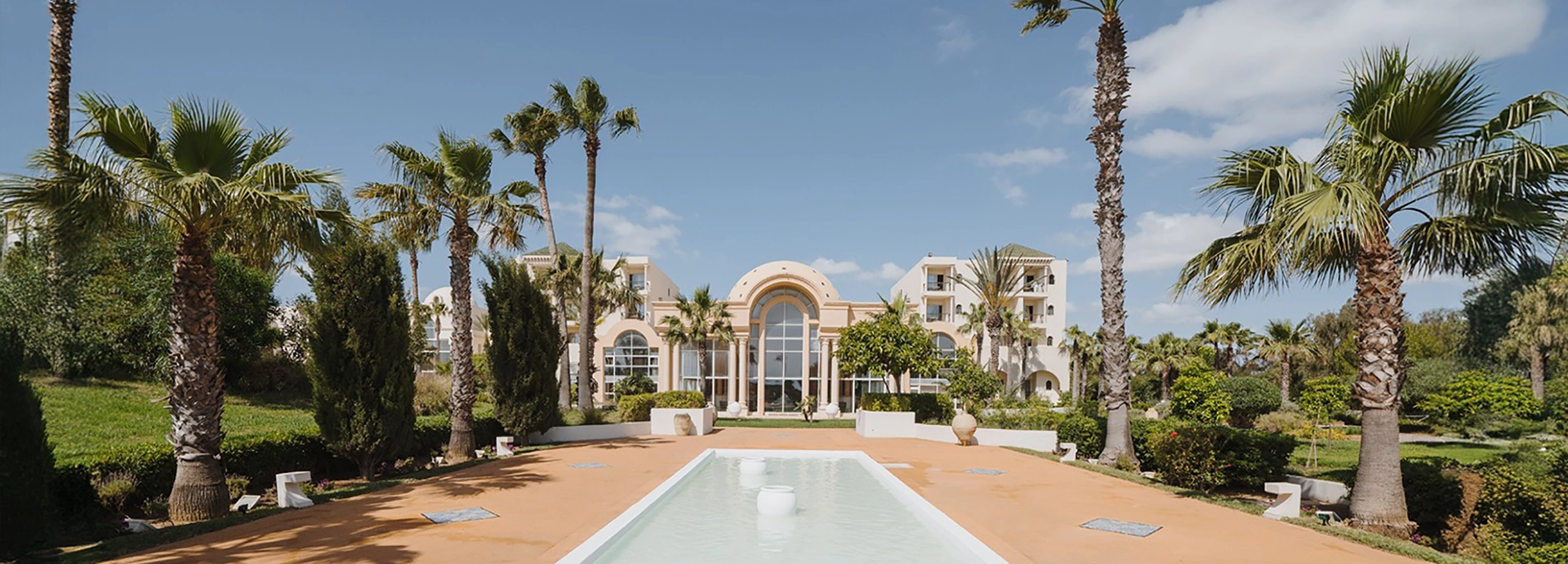 Front view of the palm-lined garden and entrance of a luxury Tunisian resort with arched windows and manicured landscaping.