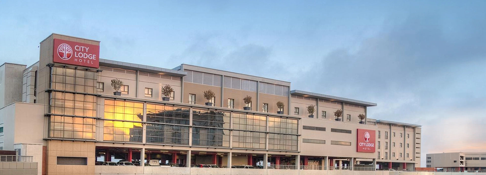 The modern, multi-story facade of the hotel glowing in the warm light of sunset, seamlessly integrated with the surrounding airport precinct.