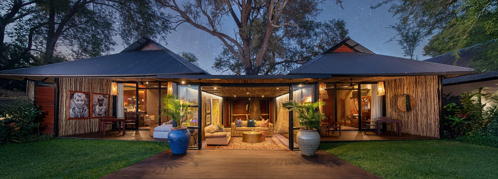 The main lodge building at Mukwa River Lodge glows under the evening sky, with open-fronted living areas connecting seamlessly to the surrounding greenery.