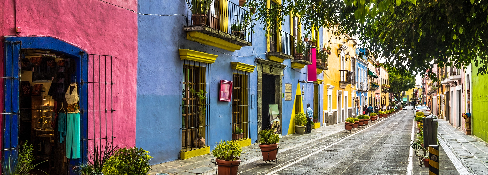A vibrant cobblestone street in Puebla lined with brightly painted colonial buildings in shades of pink, blue and yellow, potted plants at each doorway and the street receding into the distance.