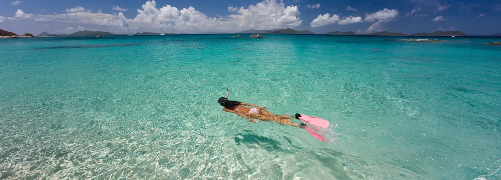 A snorkeller glides through shallow, crystal-clear turquoise water, pink fins breaking the surface, with small islands and sailing boats visible on the distant horizon under a bright blue sky.