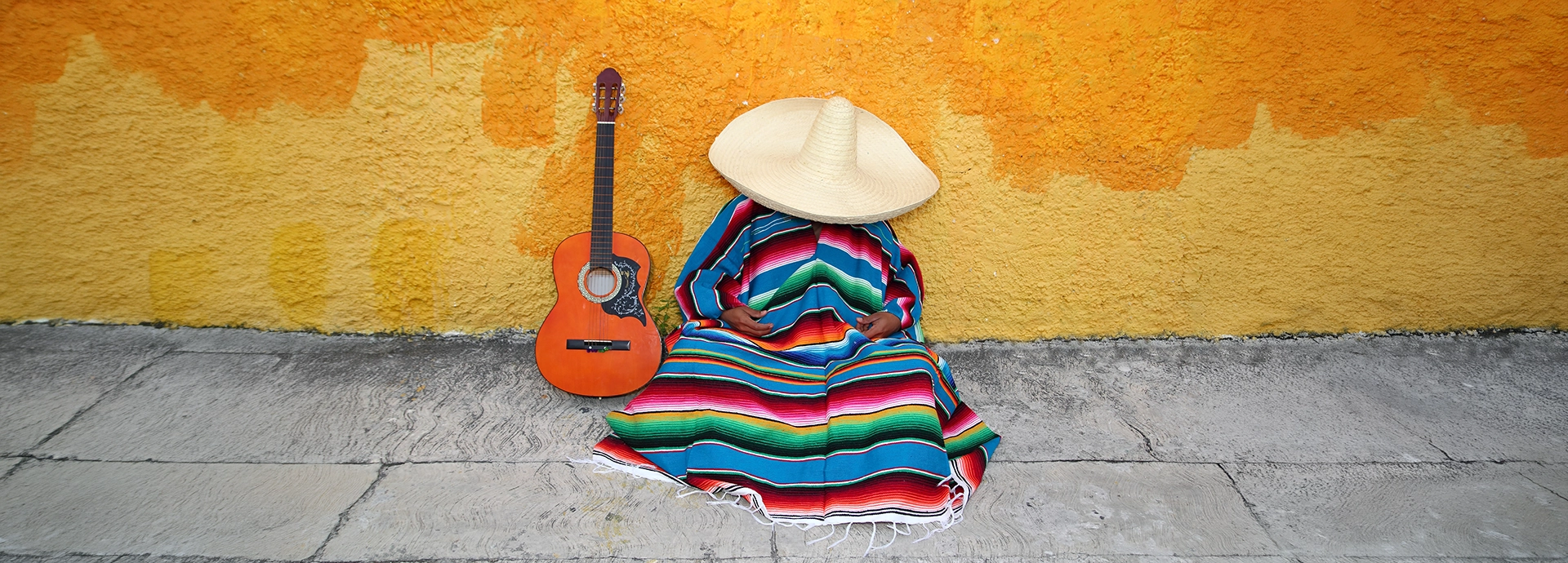 A figure wearing a colourful striped serape and wide-brimmed sombrero rests against a vivid yellow-painted wall beside an acoustic guitar on a cobblestone pavement.