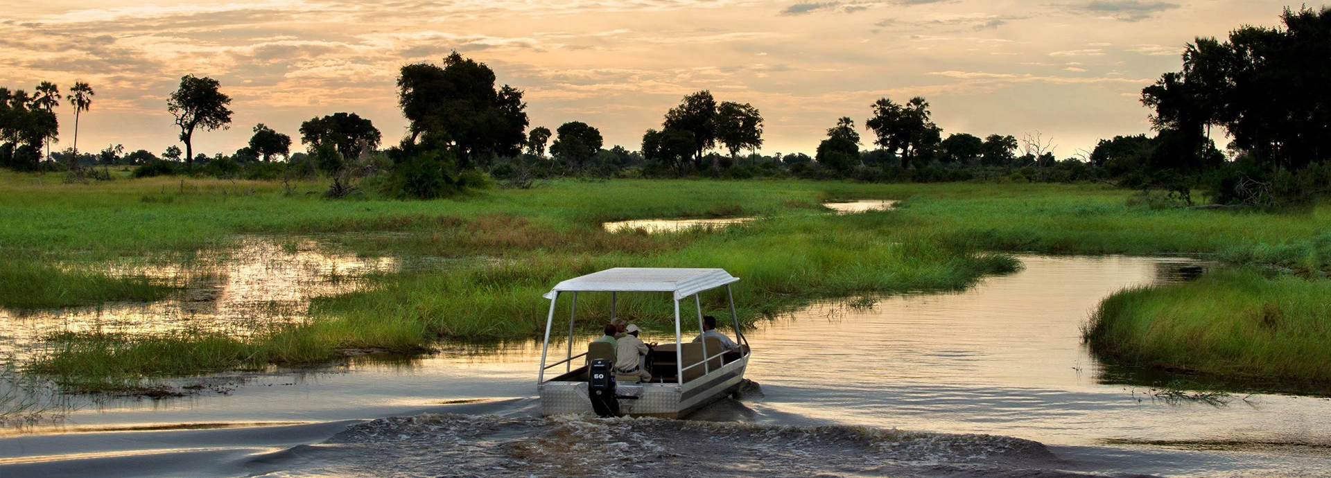 A small motorboat carrying a guide and guests moves through a flooded channel of the Okavango Delta at sunset, surrounded by wide green floodplains glowing under a golden sky.