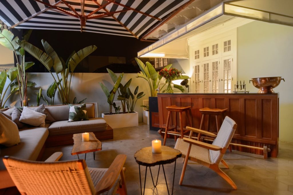 Rooftop lounge at dusk with a striped umbrella, banana palms and candlelit tables beside a dark wood bar