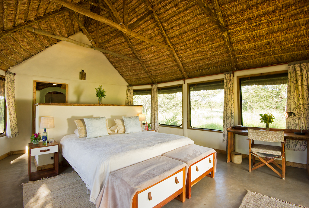 Bright thatched bedroom at El Karama Lodge with a king-size bed, linen-covered benches and wide windows onto the bush.