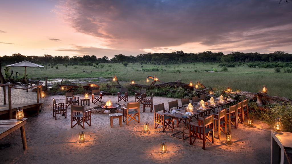 An open-air bush dinner set-up at Somalisa Bush Camp at dusk, with lantern-lit tables and director's chairs arranged in the sand around a central fire pit, overlooking a green floodplain under a dramatic pink and purple sky, Hwange National Park, Zimbabwe.